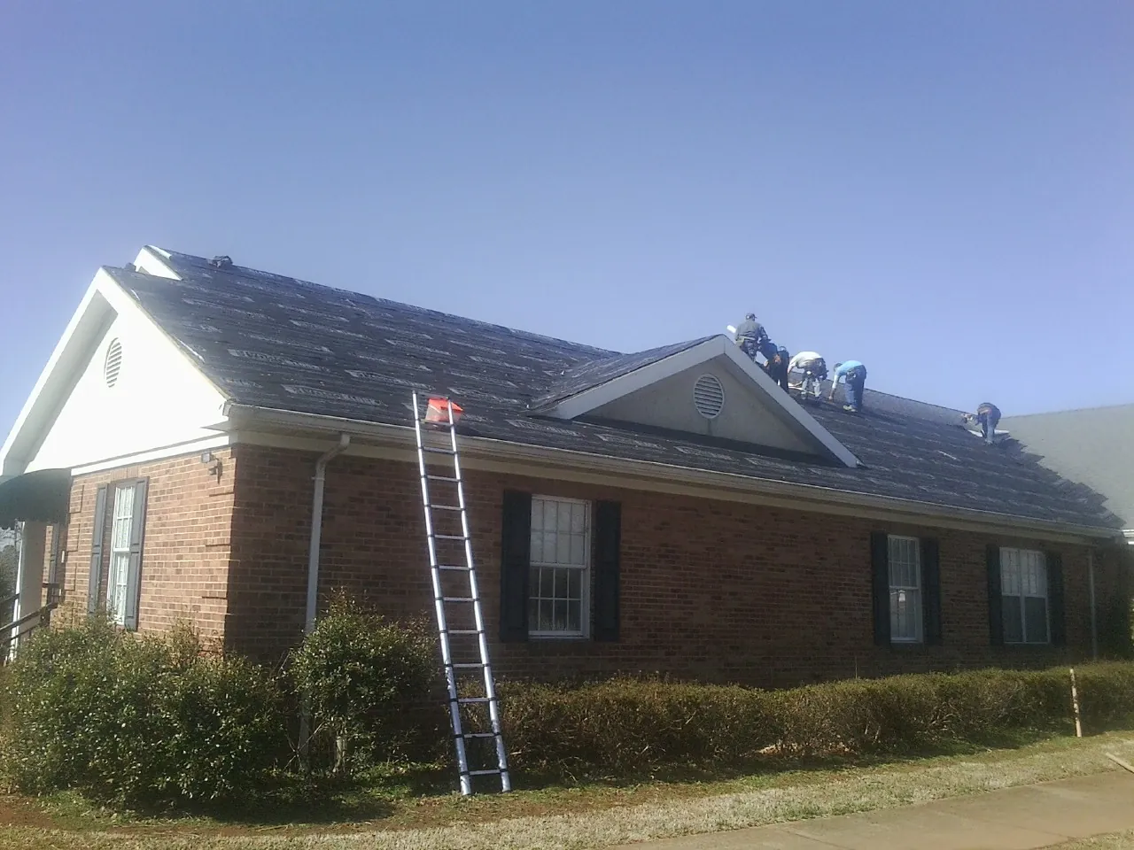 Crew tearing shingles off a brick ranch roof with a ladder set — Emergency Roof Repair work by our crew in Lafitte